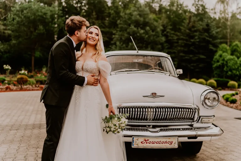 Pan młody całuje pannę młodą obok zabytkowego samochodu – fotograf ślubny Łódź